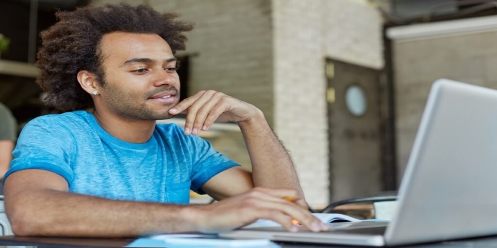 Pessoa escrevendo em um caderno enquanto utiliza um laptop sobre uma mesa de estudo