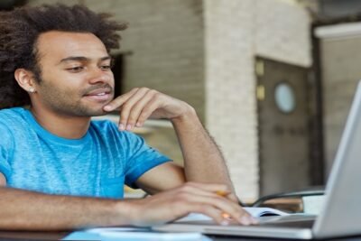Pessoa escrevendo em um caderno enquanto utiliza um laptop sobre uma mesa de estudo