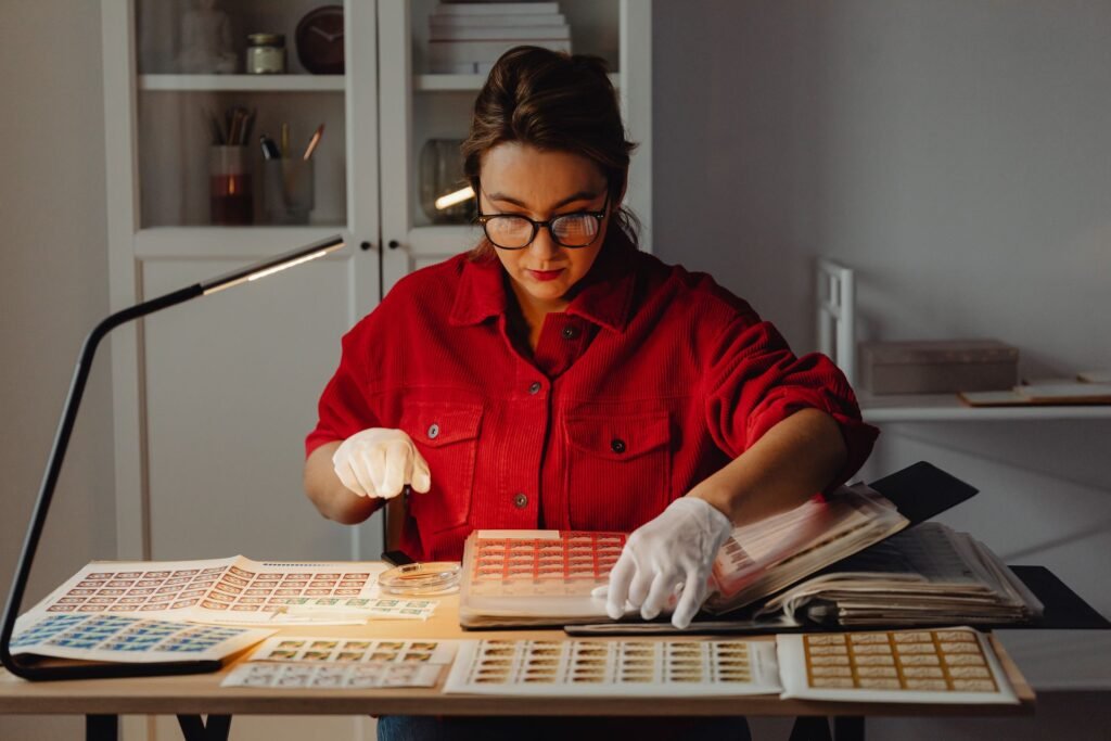 Mulher de camisa vermelha usa luvas brancas para organizar folhas de selos e álbuns sobre uma mesa iluminada por luminária, em ambiente doméstico