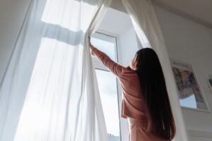Mulher de costas abre cortinas brancas e deixa a luz natural entrar no quarto pela manhã