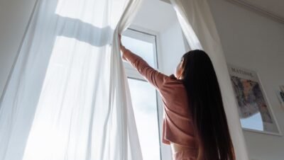 Mulher de costas abre cortinas brancas e deixa a luz natural entrar no quarto pela manhã