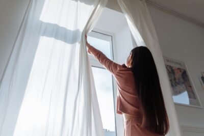 Mulher de costas abre cortinas brancas e deixa a luz natural entrar no quarto pela manhã